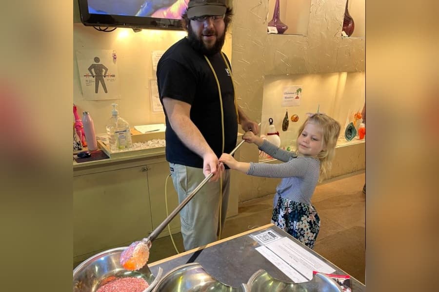 Instructor helps a young girl hold a glassblowing rod with molten glass at a glass studio workshop.