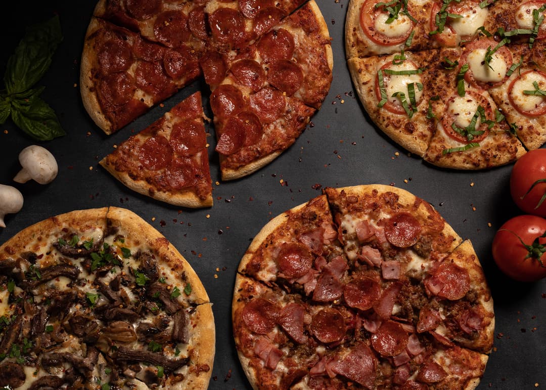 Four assorted pizzas on a dark surface with fresh basil, mushrooms, and tomatoes nearby.