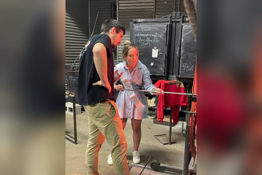Two people in a glassblowing studio working together with a metal rod near furnaces and equipment.