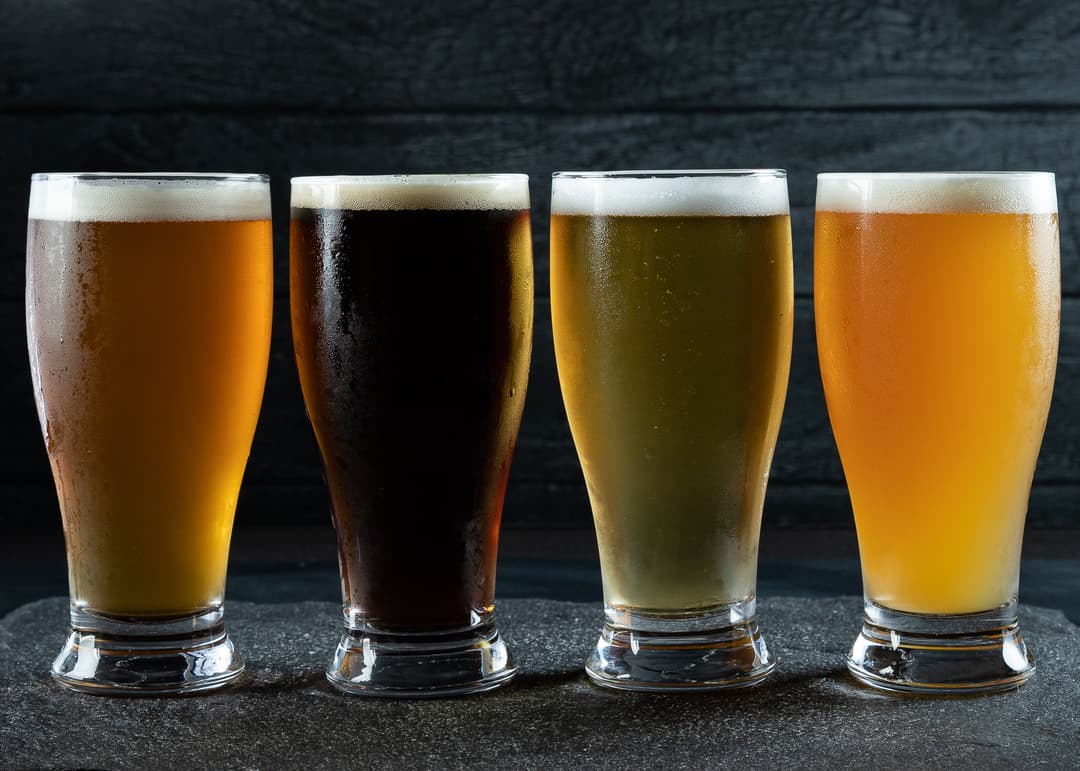 Four glasses of beer in varying colors from amber to dark to pale, against a dark background.