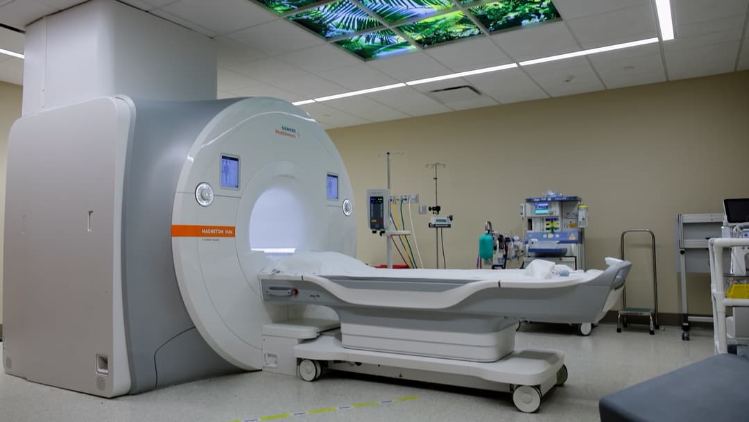 Siemens MRI scanner in a hospital room with anesthesia equipment and a backlit ceiling panel of green ferns.