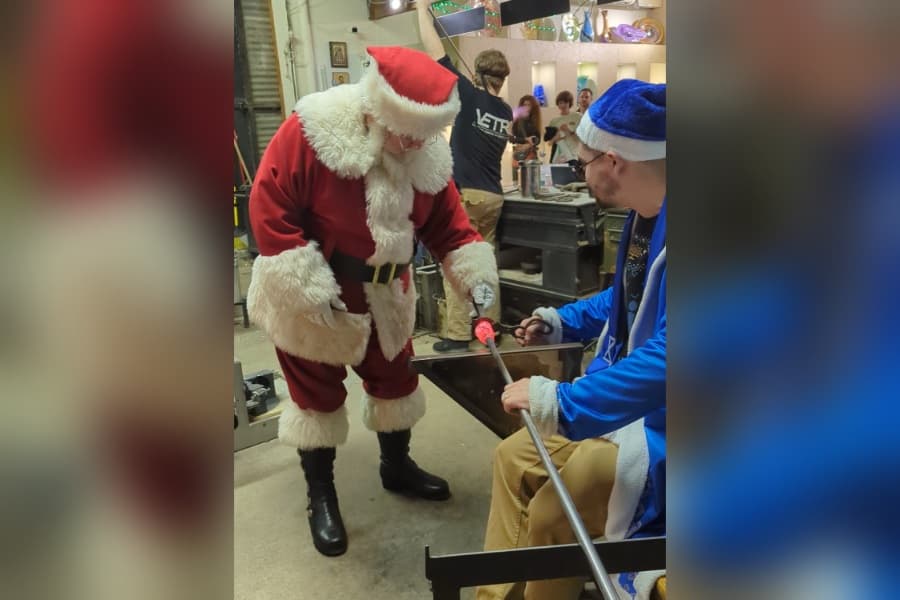Santa Claus works molten glass with a tool while a man in a blue hat holds the blowpipe in a glass studio.