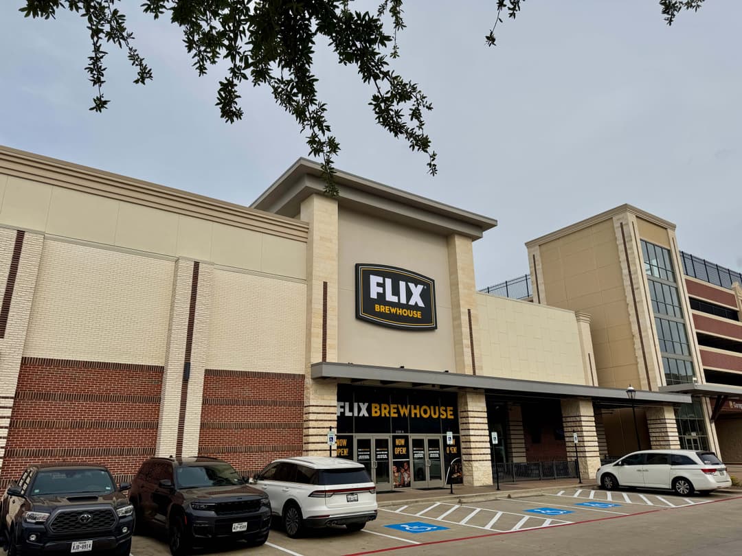 Exterior of Flix Brewhouse theater with "Now Open" signs and cars parked in the lot under a cloudy sky.