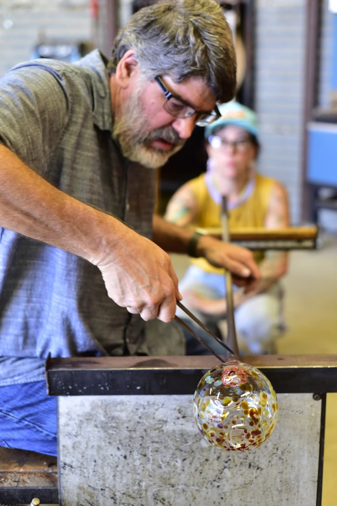A glassblower shapes a speckled glass sphere with tools as a woman watches in the background of a studio.
