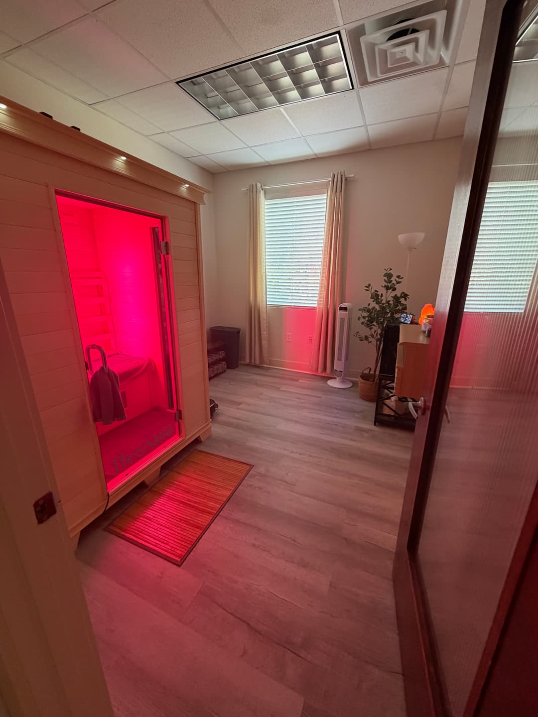 Wellness room with a red-lit infrared sauna, wood floors, window, plant, fan, and salt lamp.