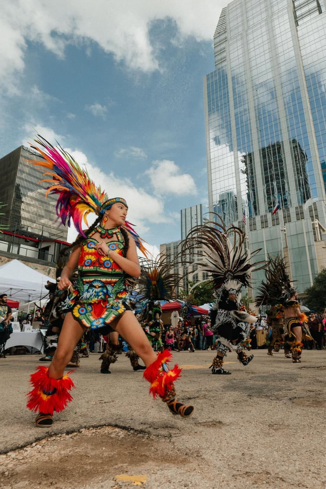 Aztec dancers in colorful feathered headdresses perform at a downtown street festival near glass skyscrapers.
