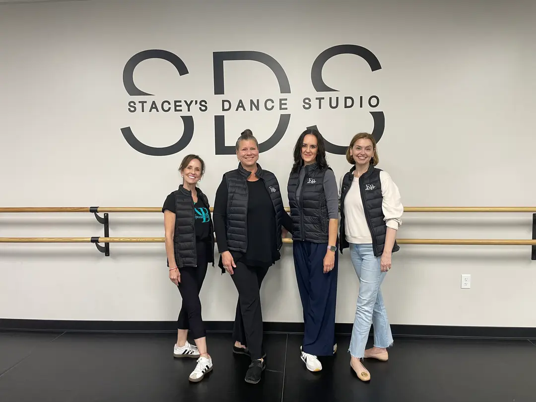 Four women in black vests stand smiling in front of a Stacey's Dance Studio wall logo.