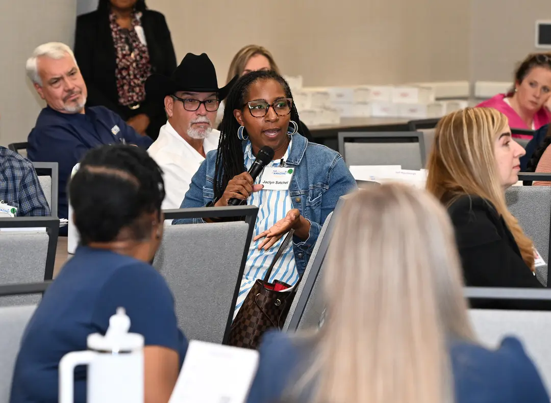 Woman in denim jacket speaks into a microphone among seated attendees at a conference.