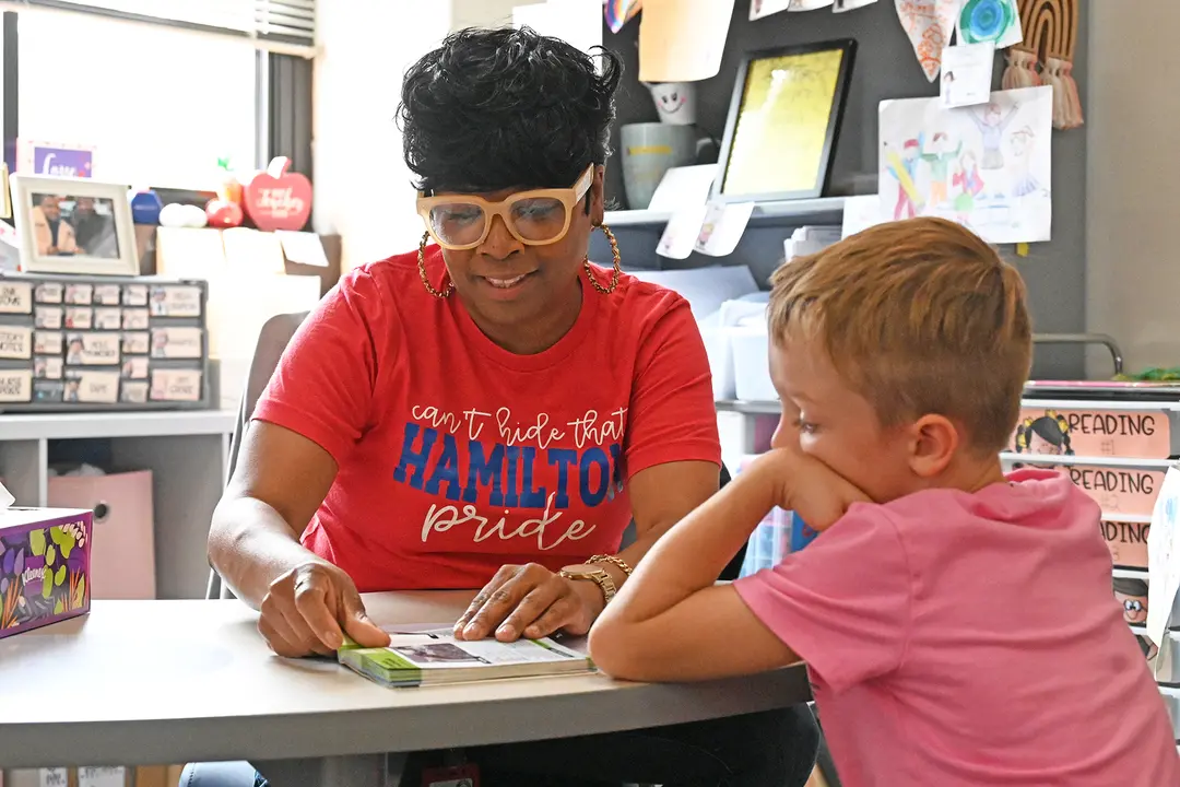 Teacher in red "Hamilton Pride" shirt reads a book with a young boy at a classroom table.