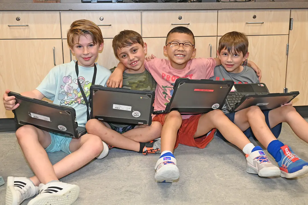 Four smiling boys sit on a classroom floor with Chromebook laptops, arms around each other.