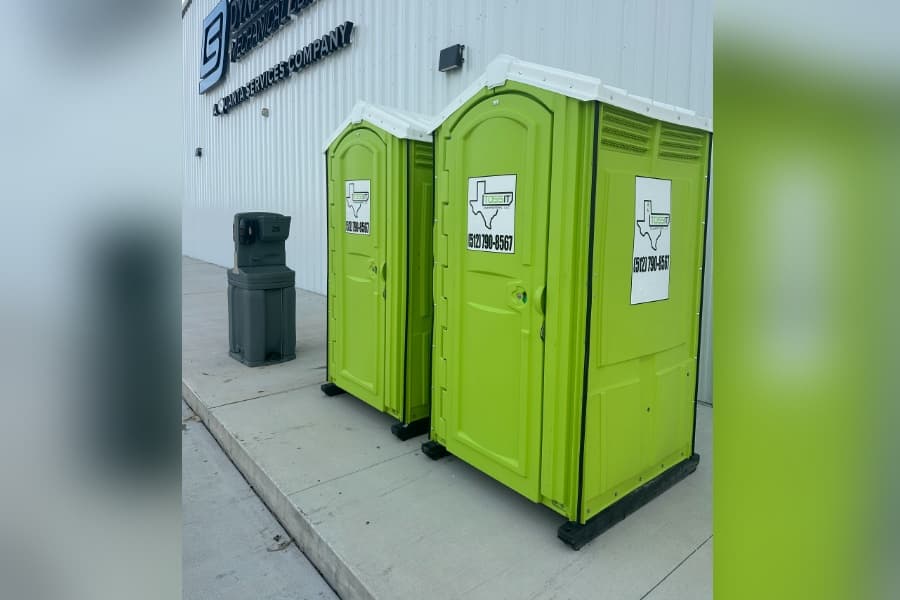 Two green portable toilets and a handwashing station outside a white building with a business sign.