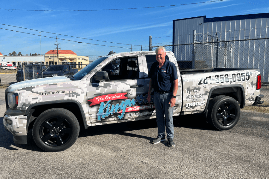 Man in navy shirt stands beside camouflage-wrapped pickup truck with air conditioning company branding and phone number.