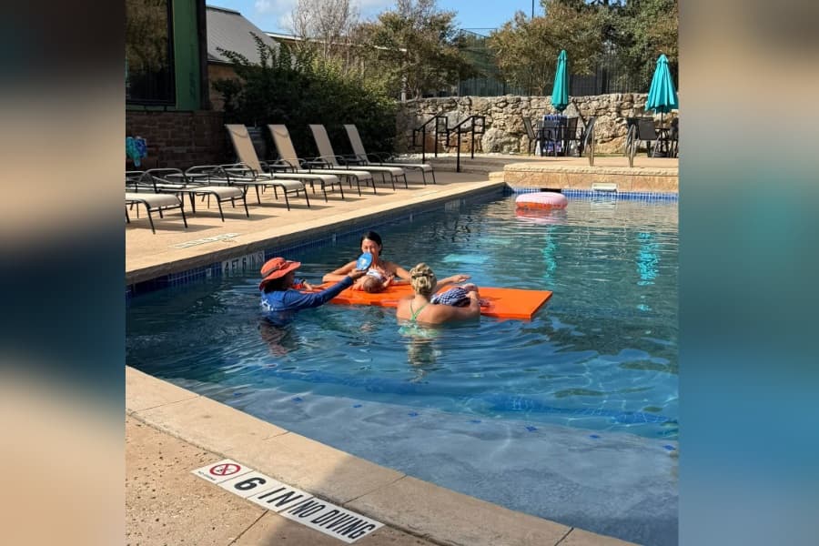 Adults in an outdoor pool holding babies on an orange float, with lounge chairs and umbrellas nearby.