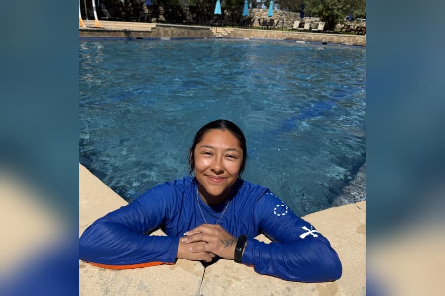 Smiling young woman in a blue shirt rests arms on the edge of an outdoor swimming pool.