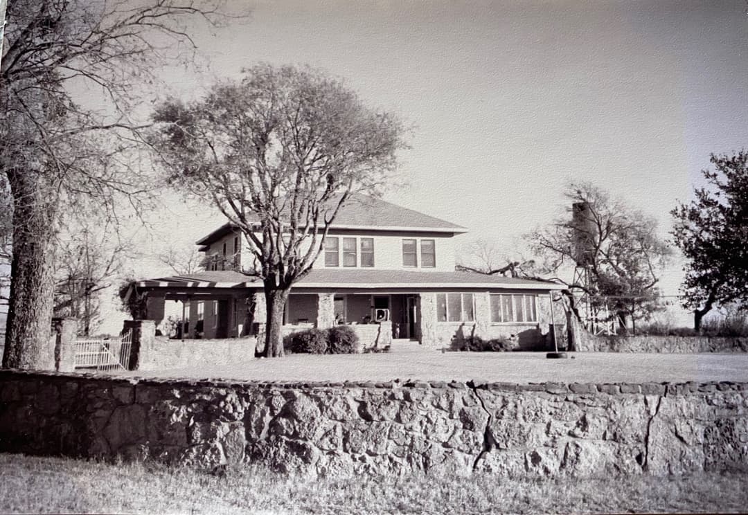 Vintage black-and-white photo of a two-story farmhouse with a stone wall, gate, and trees in the yard