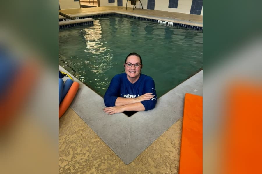 Smiling woman in blue shirt and glasses leans on edge of indoor pool, standing in the water.