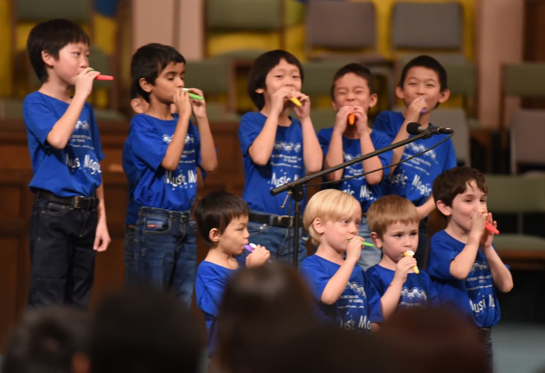 Boys in matching blue "Music Magic" shirts play colorful whistles on stage near a microphone.