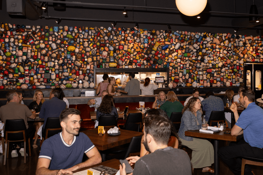 Busy restaurant with diners at tables, an open kitchen, and a wall covered in colorful decorative objects.