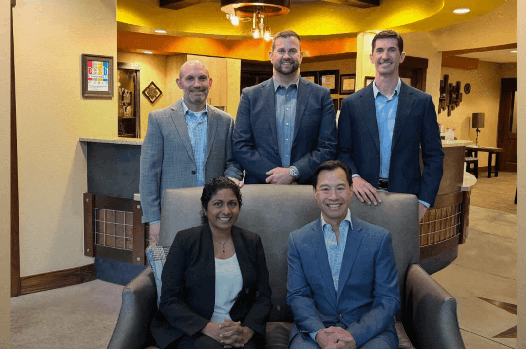 Five professionals in business attire pose smiling in an office lobby, two seated and three standing.