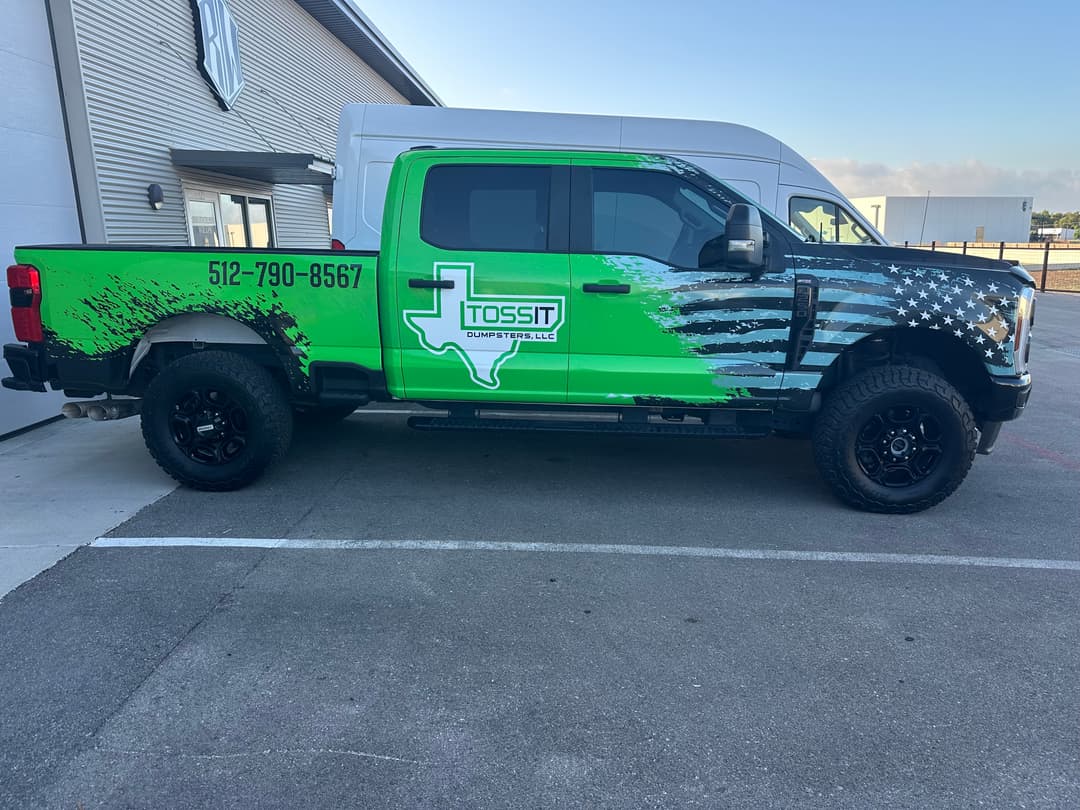 Green pickup truck wrapped with Toss It Dumpsters LLC logo and phone number 512-790-8567, parked outside a building.