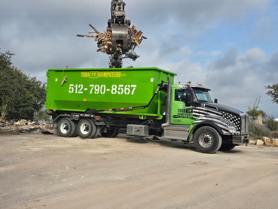 Excavator grapple drops debris into green Toss It Dumpsters roll-off truck with flag design on cab.