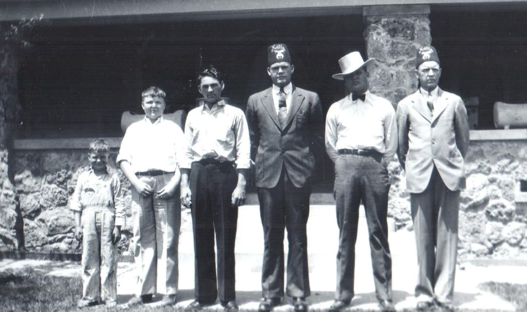 Four men, two wearing Shriner fezzes, and two boys stand before a stone building in a vintage black-and-white photo.