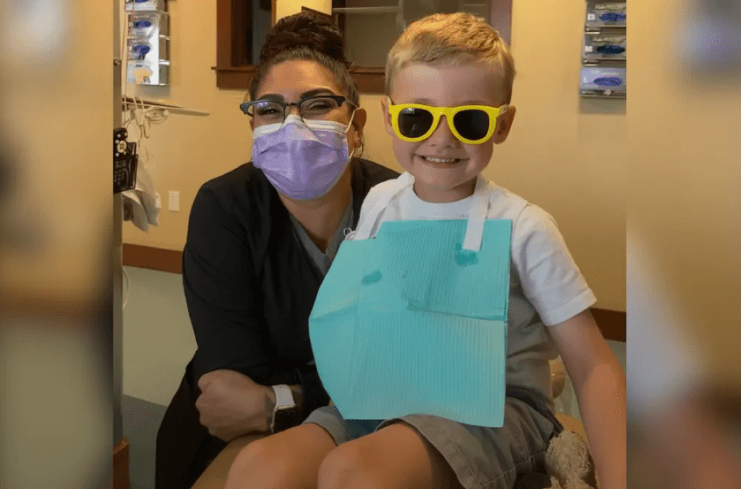 Smiling boy in yellow sunglasses with dental bib beside a masked dental worker in a dentist's office.