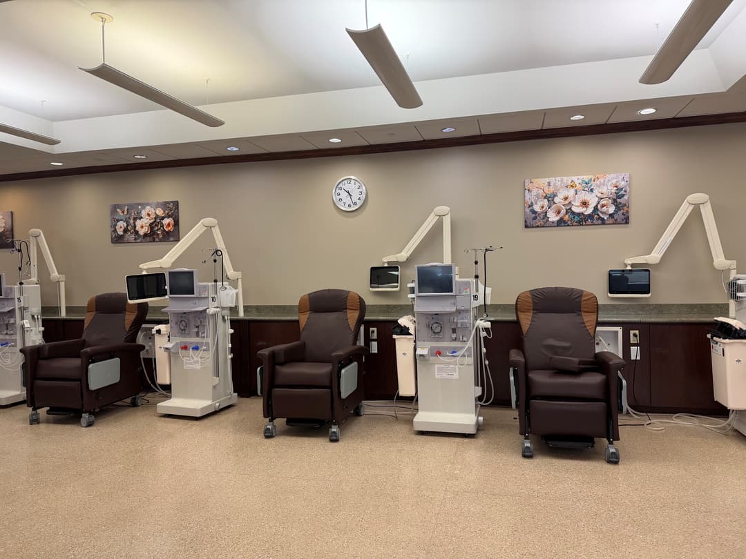 Dialysis treatment room with brown recliner chairs, dialysis machines, wall clock and floral artwork.