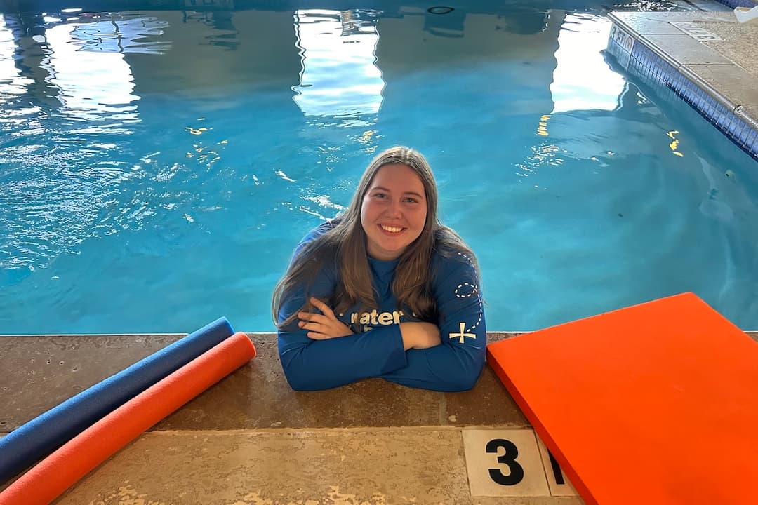 Smiling woman in blue swim shirt leans on pool edge beside foam noodles and a kickboard.