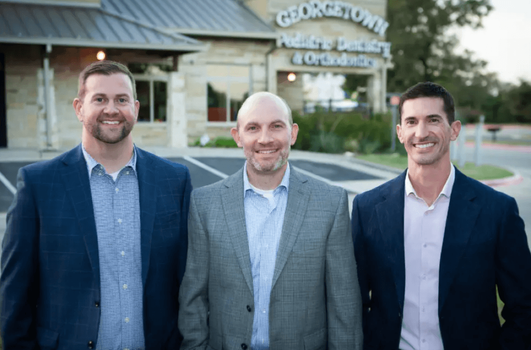 Three smiling men in suits stand outside Georgetown Pediatric Dentistry & Orthodontics building.