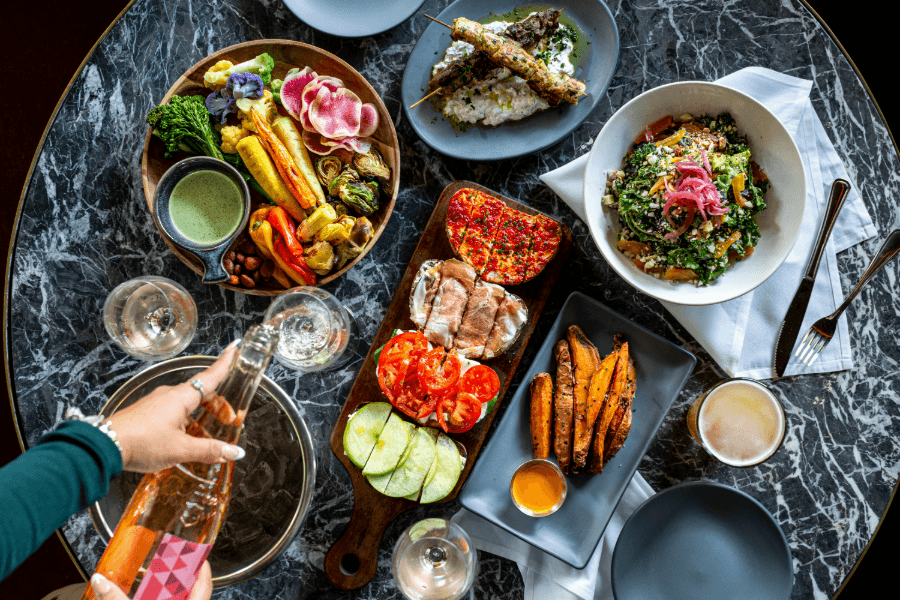 Overhead view of a marble table with roasted vegetables, salad, fries, skewers, and a hand pouring rosé wine.