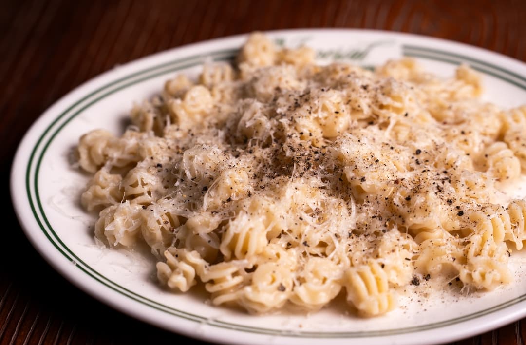 Pasta topped with grated cheese and black pepper on a green-rimmed plate atop a dark wooden table.