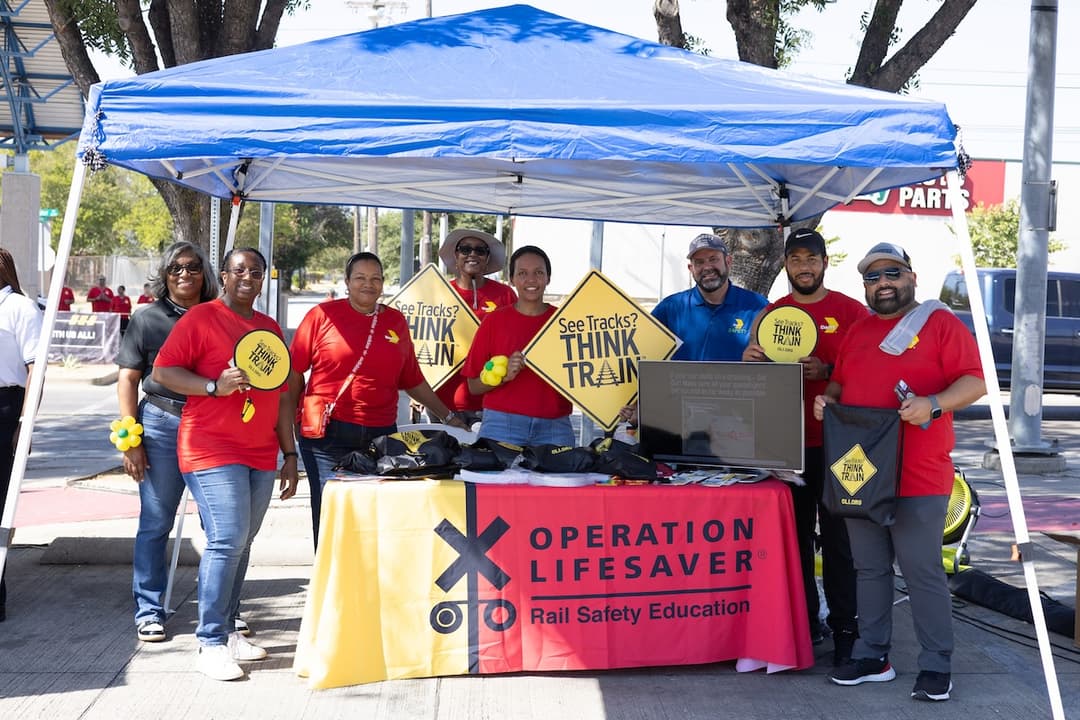 Group at Operation Lifesaver rail safety booth under blue tent, holding "See Tracks? Think Train" signs.