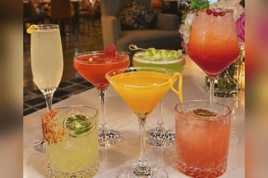 Seven colorful cocktails in various glasses with garnishes displayed on a marble table.