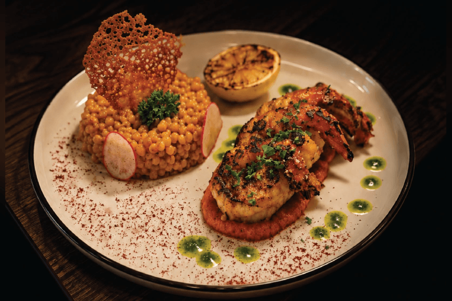 Grilled shrimp on red sauce with couscous, grilled lemon, radish, and herb oil on a plated dish.