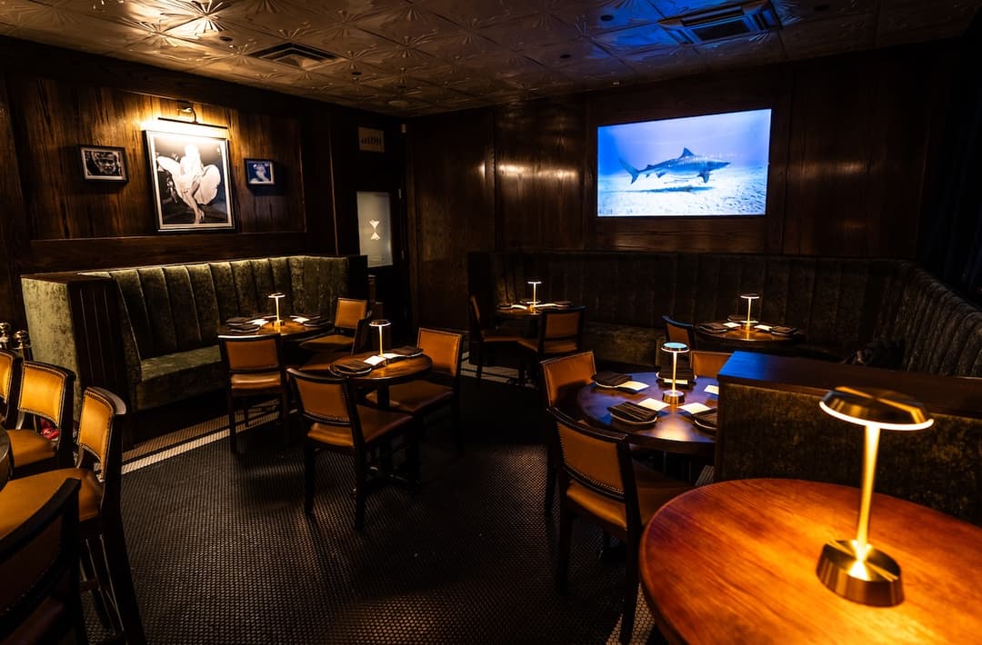 Dimly lit restaurant with wood paneling, leather chairs, green banquettes, table lamps, and a wall screen showing a shark.