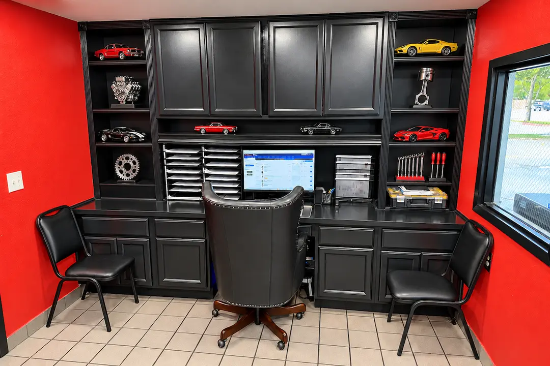 Automotive office with black cabinetry, red walls, desk with monitor, leather chair, and car models on shelves.