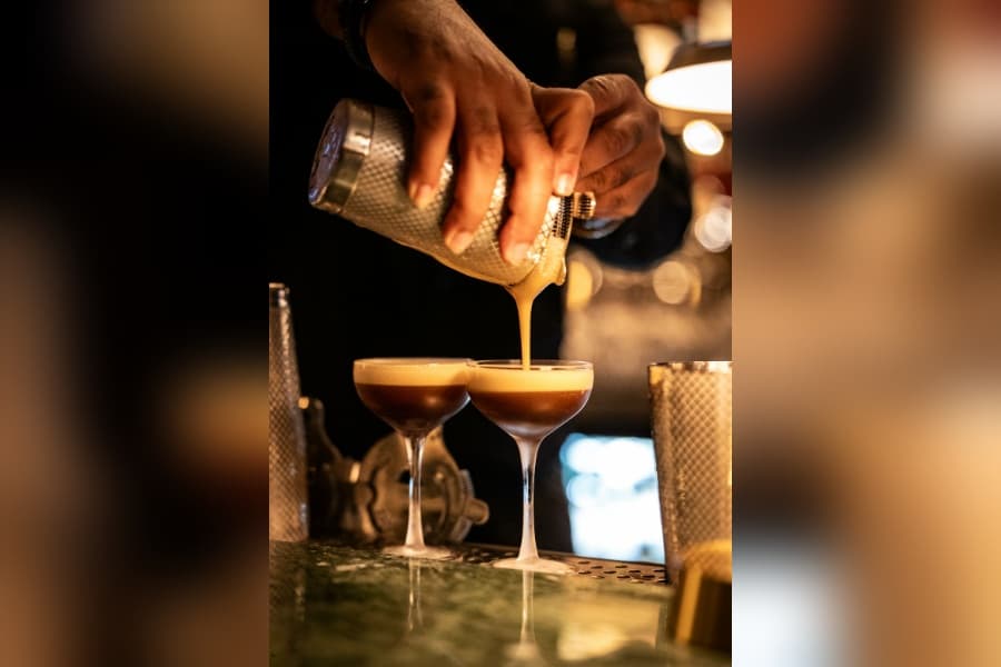 Bartender pours an espresso martini from a shaker into two coupe glasses at a dimly lit bar.