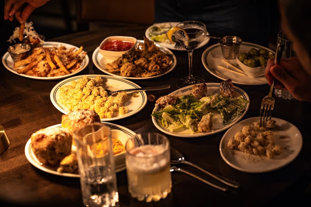 Dimly lit table spread with fries, pasta, Caesar salad, cocktails and martini as diners share small plates.