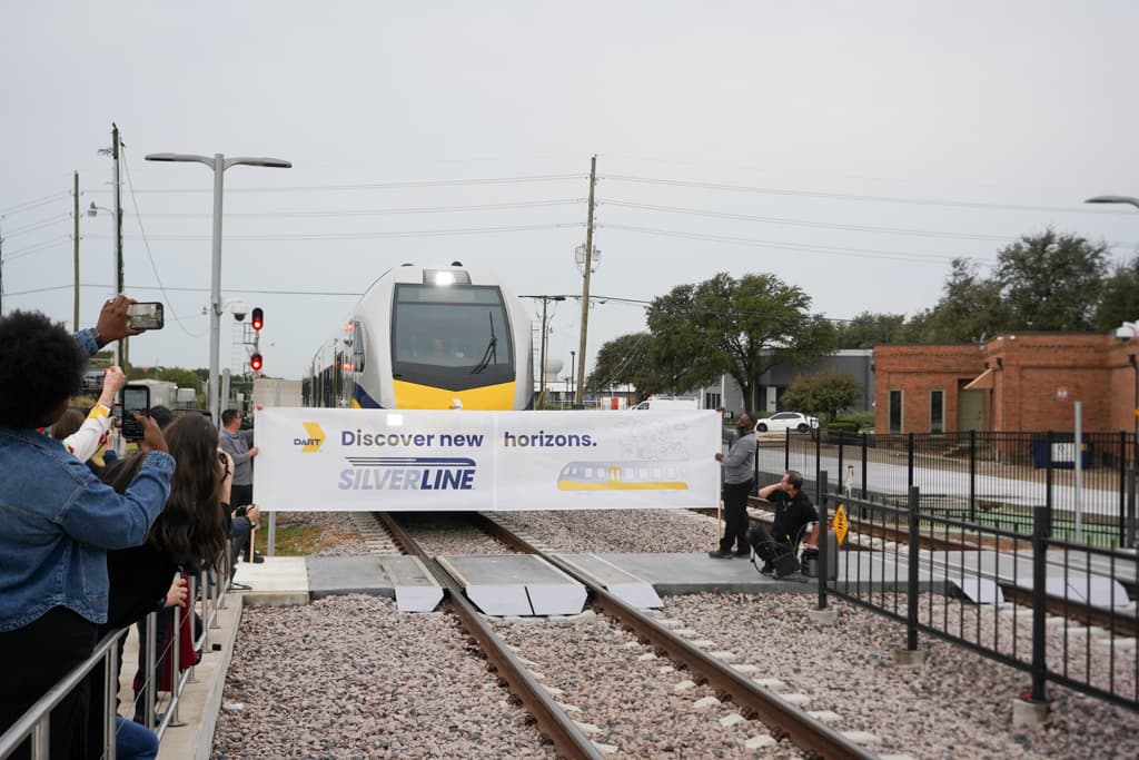 DART Silverline train approaches a banner reading "Discover new horizons" as spectators watch and photograph.