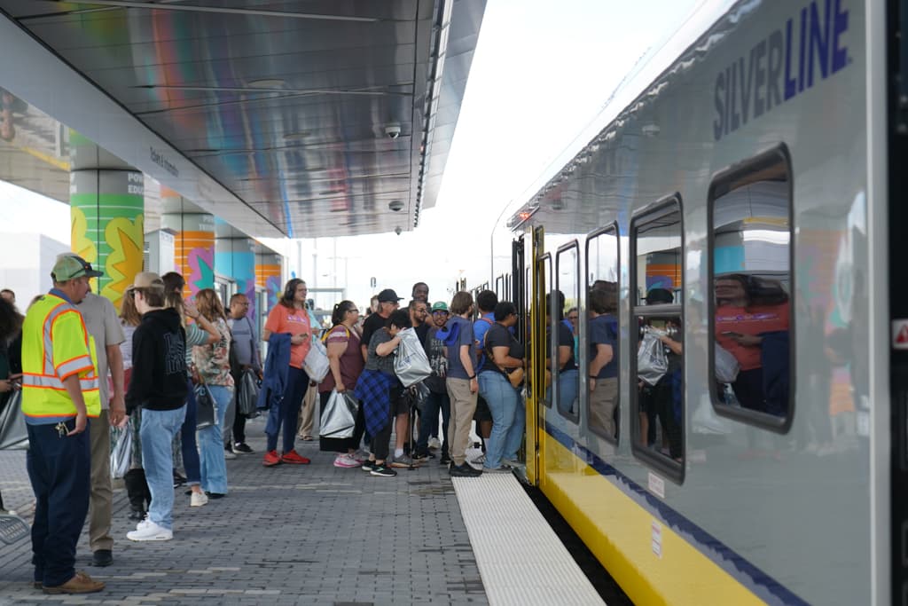 Crowd of people boarding a Silver Line train at a station platform, with a worker in a yellow safety vest nearby.
