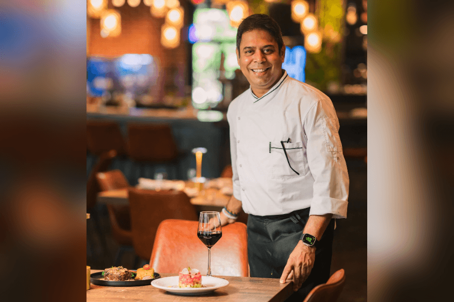 Smiling chef in white jacket stands by a restaurant table set with plated dishes and a glass of red wine.