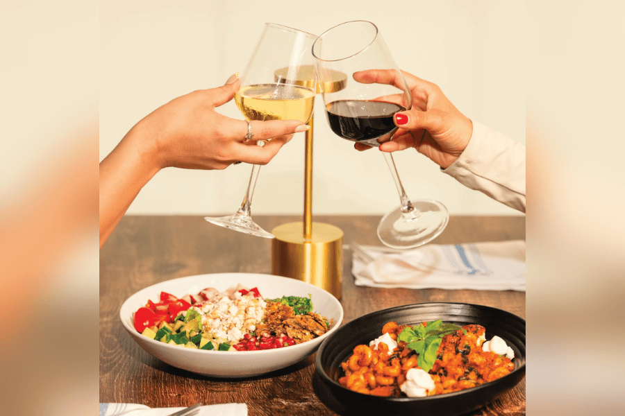 Two hands toast glasses of white and red wine over a salad and pasta dish on a wooden table.