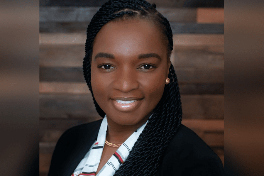 Smiling woman with braided hair wearing a black blazer and patterned blouse against a wood-panel background.