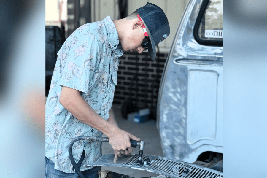Young man in floral shirt and cap uses a pneumatic tool to work on a vehicle's chrome grille.