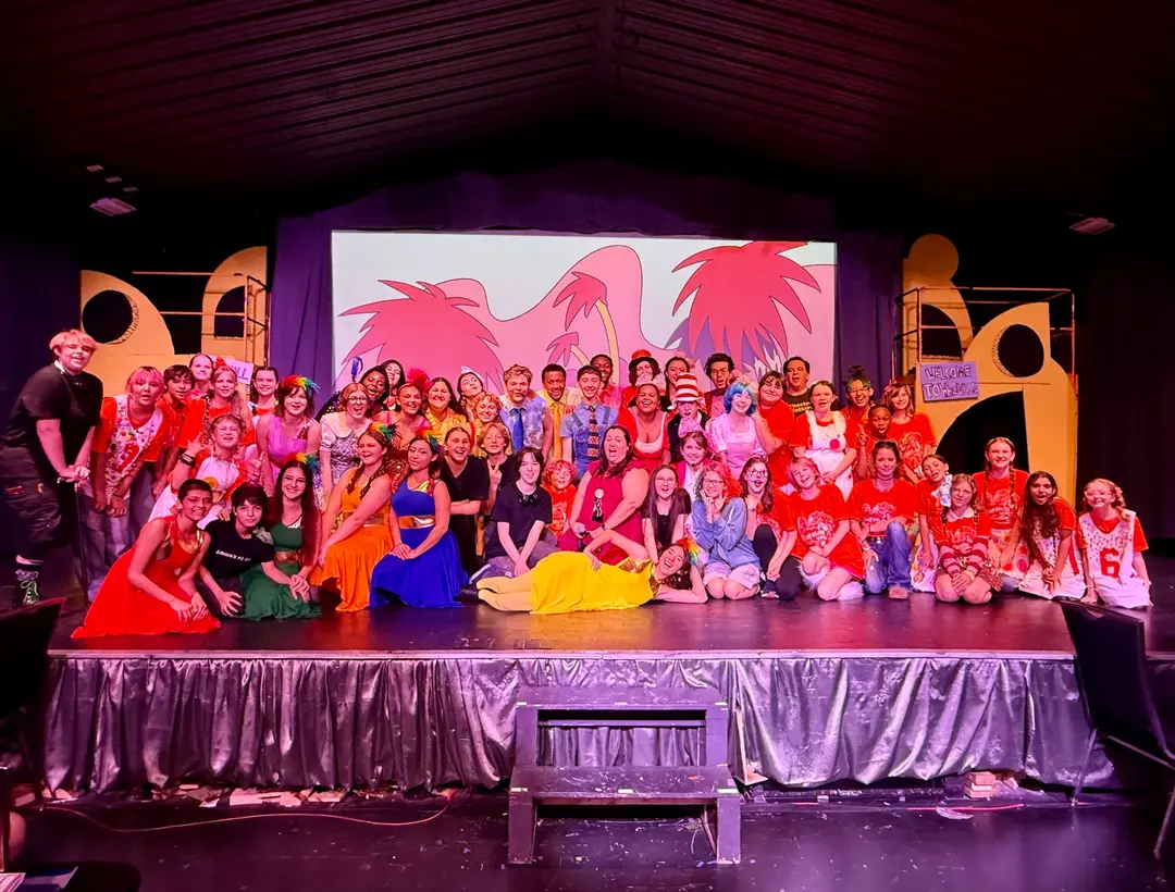 Large cast of a Seussical theater production poses on stage in colorful costumes before a palm tree backdrop.