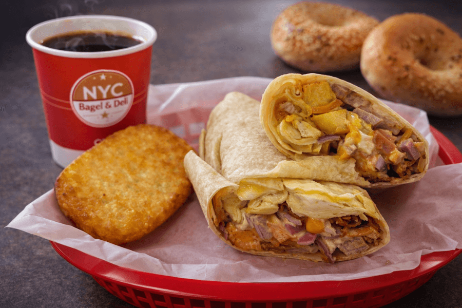 Breakfast burrito and hash brown in a red basket beside a steaming NYC Bagel & Deli coffee cup and two bagels.