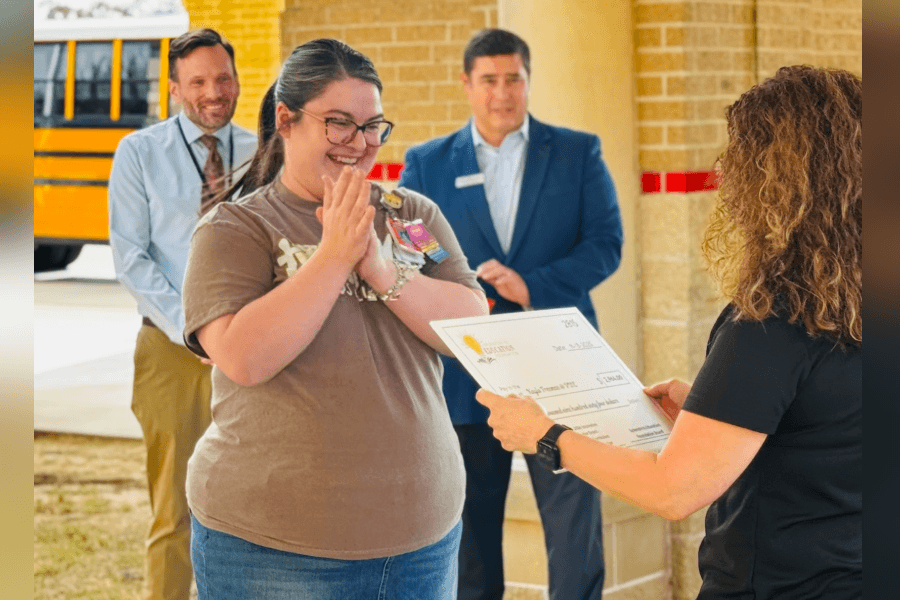 Smiling woman reacts with hands clasped as she receives a large check, with two men and a school bus behind.