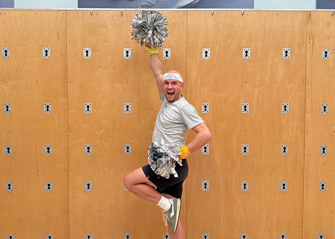 Smiling man in headband and shorts jumps with silver pom-poms raised against a wooden wall.