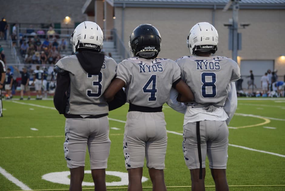 Three NYOS football players in gray jerseys numbered 3, 4, and 8 stand arm-in-arm on the field.
